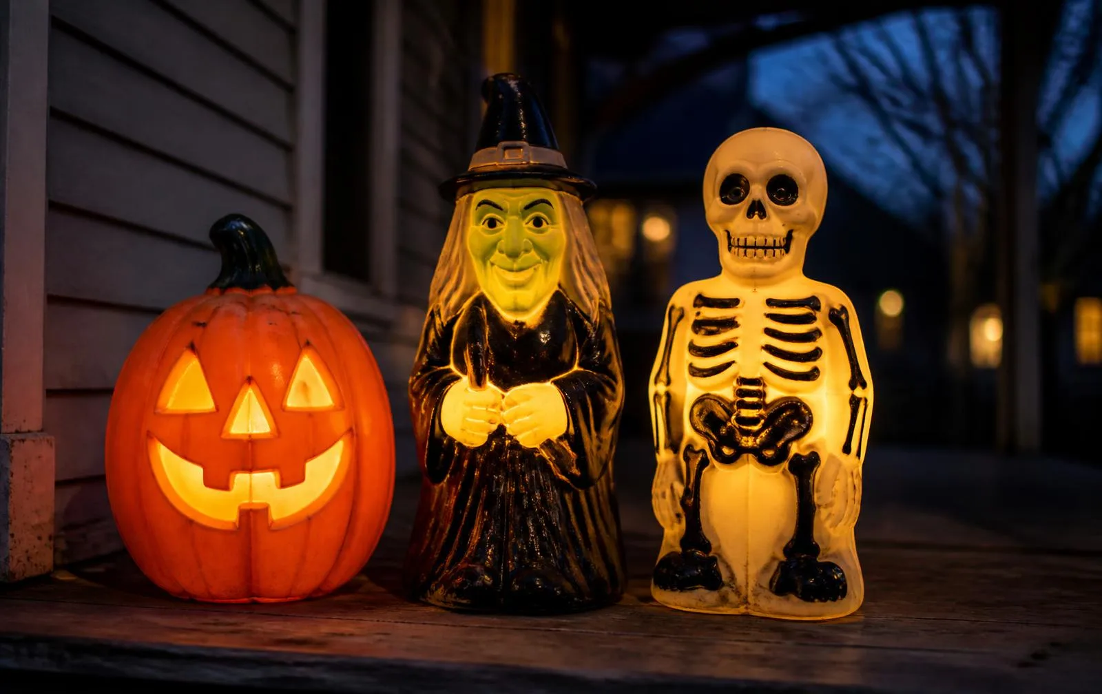 Three vintage Halloween blow molds lit on a porch at dusk: an orange jack-o'-lantern, a green-faced witch, and a skeleton
