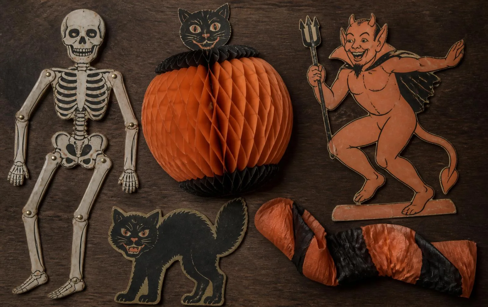 Flat lay of 1950s die-cut cardboard Halloween decorations: a jointed skeleton, an orange honeycomb pumpkin centerpiece, black cat cutouts and a crepe paper streamer