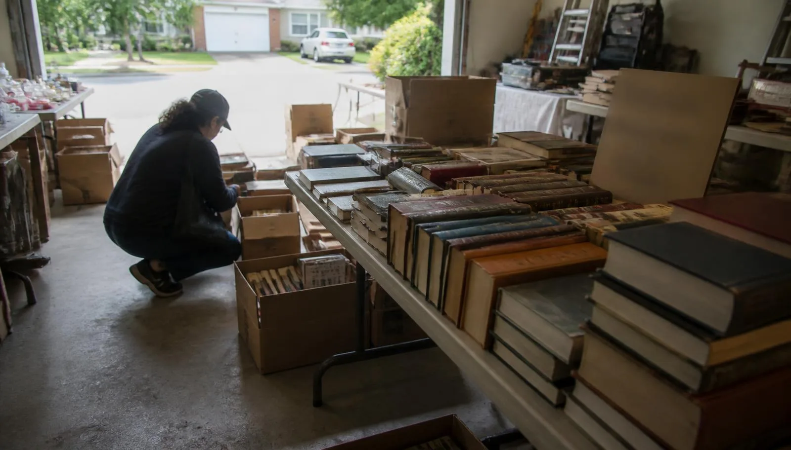 A shopper crouching to dig through cardboard boxes of old hardcover books at a garage estate sale