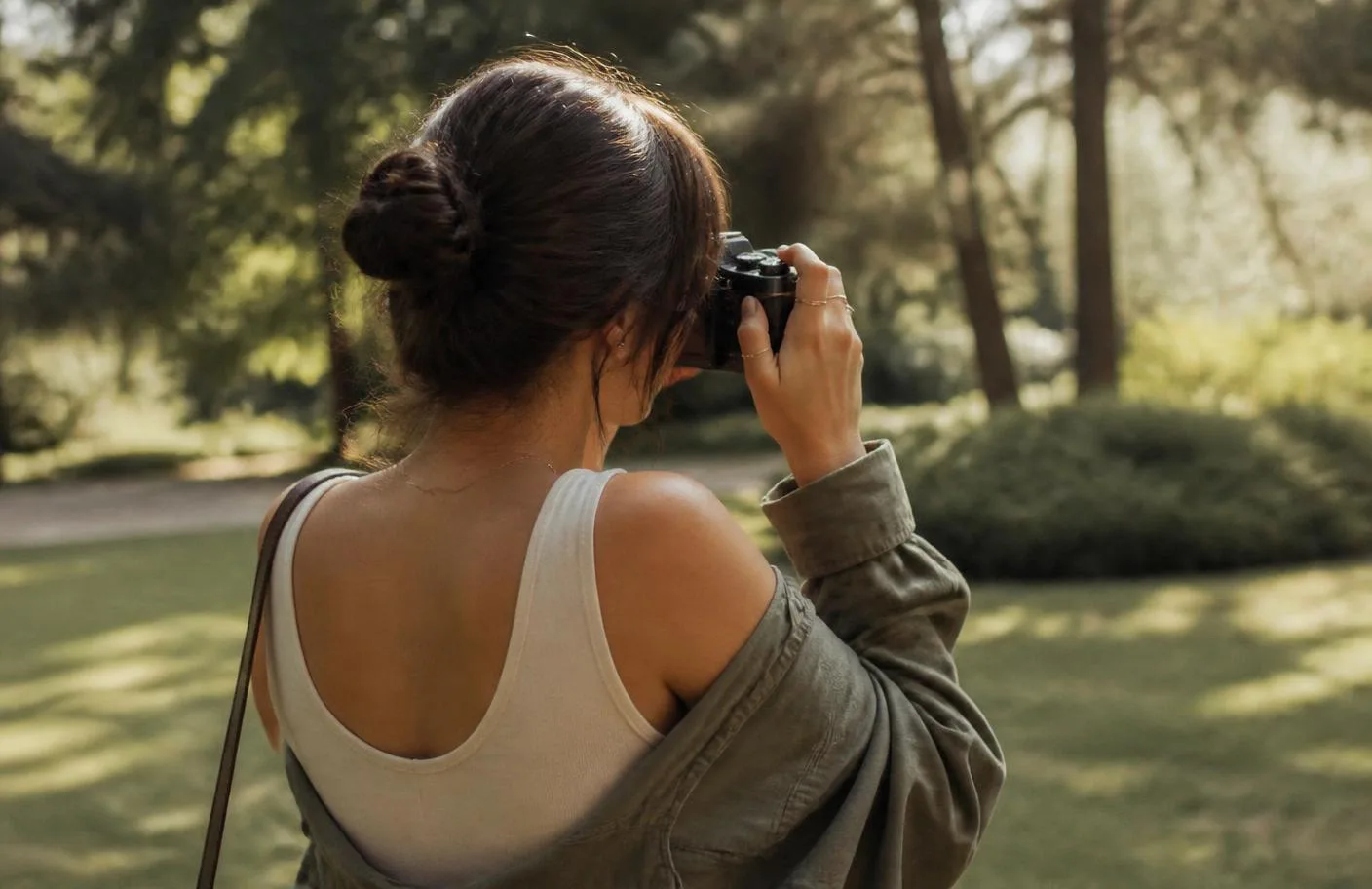 A young woman photographed from behind holding a black Pentax K1000 up to her eye in a sunlit park, wearing a thrifted-vintage outfit