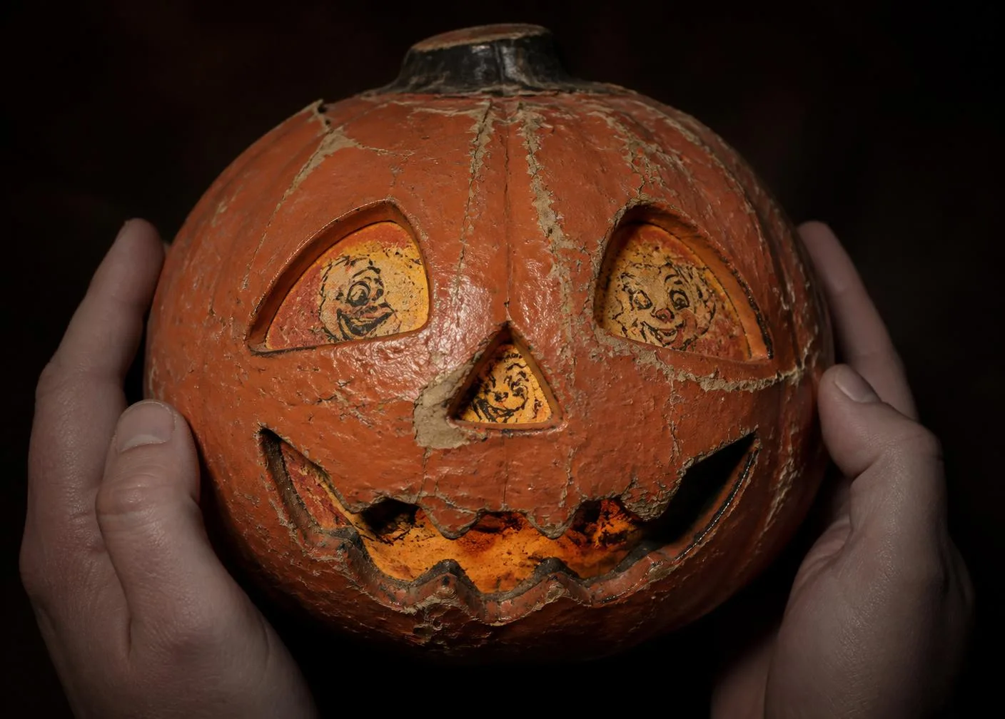 Close-up of a 1930s papier-mache jack-o'-lantern held in two hands, showing crazed paint and a glowing printed paper face insert