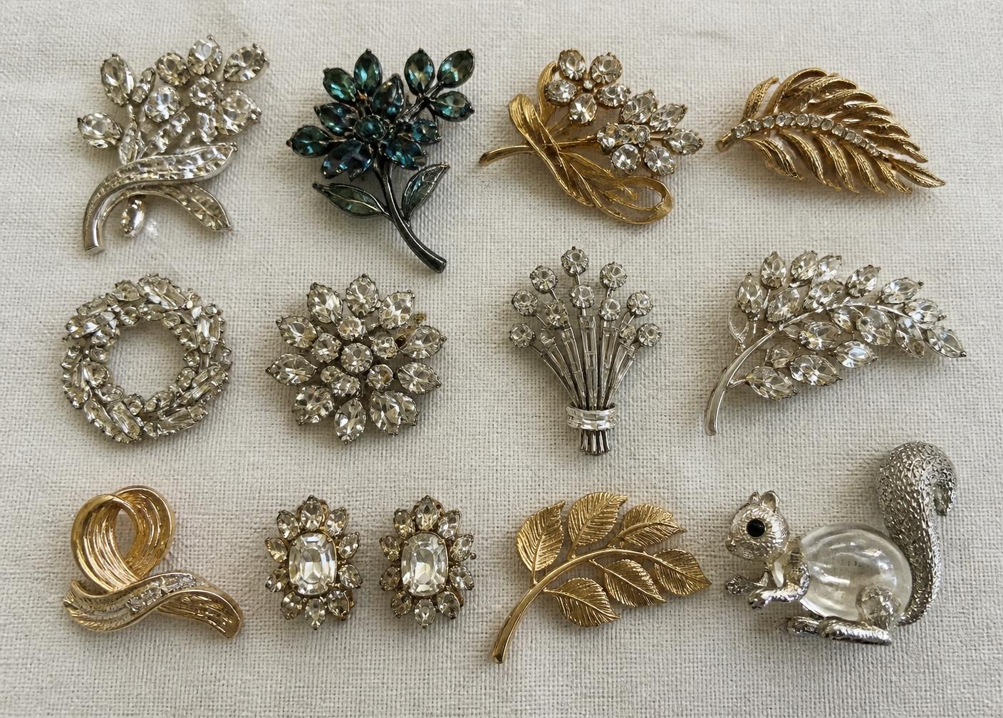 Fourteen vintage rhinestone and gold-tone brooches and clip earrings arranged in rows on cream linen, including a japanned black floral pin and a clear Lucite figural animal pin