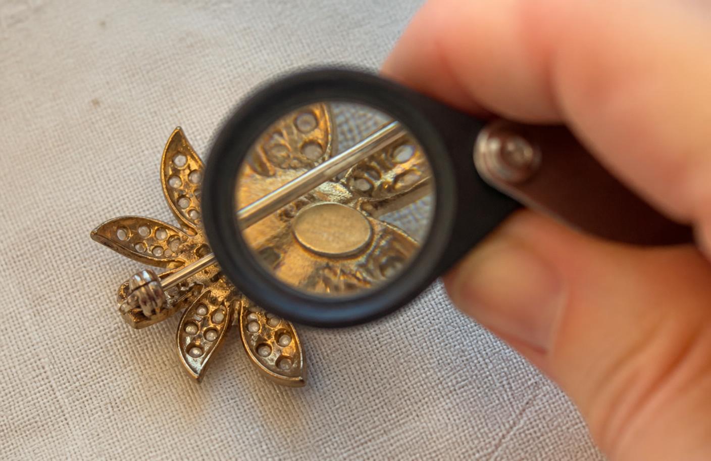 A hand holding a jeweler's loupe over the back of a vintage rhinestone flower brooch, showing the pin stem, safety catch and a small stamped oval maker's plaque