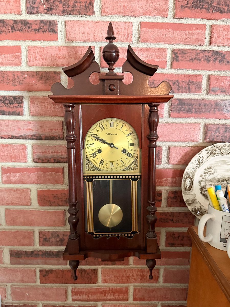 An ornate mahogany pendulum wall clock with a brass Roman-numeral face hanging on a brick wall