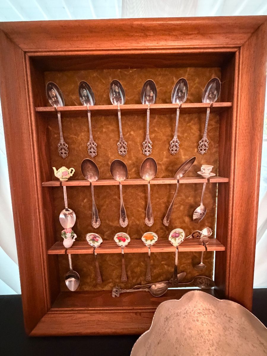 A framed wooden display case holding a collection of vintage souvenir and enamel-handled spoons