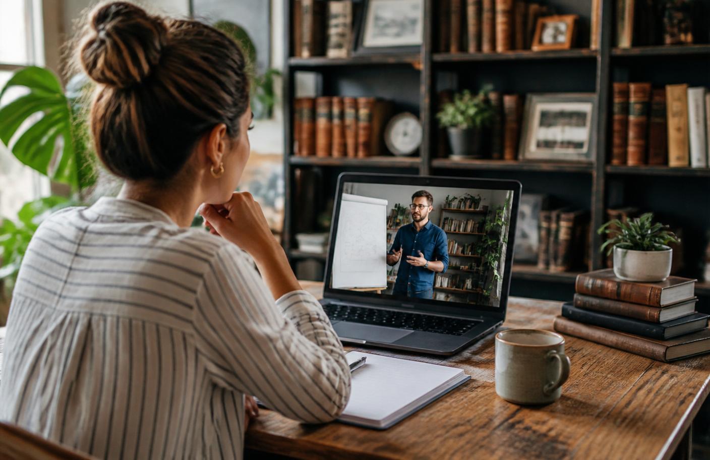 A woman entrepreneur watching a business training on her laptop in a cozy home office with plants and vintage books — sharpening the craft.