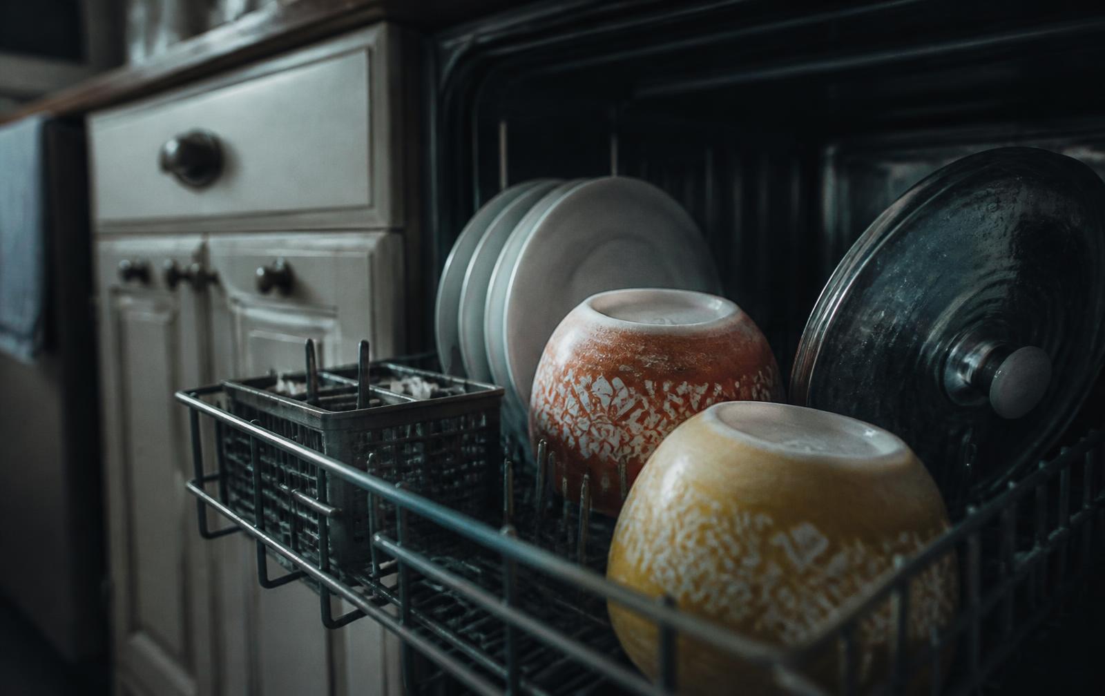 Vintage patterned Pyrex bowls loaded into a modern dishwasher rack among white plates, with a faint mineral film visible on the vintage pieces.