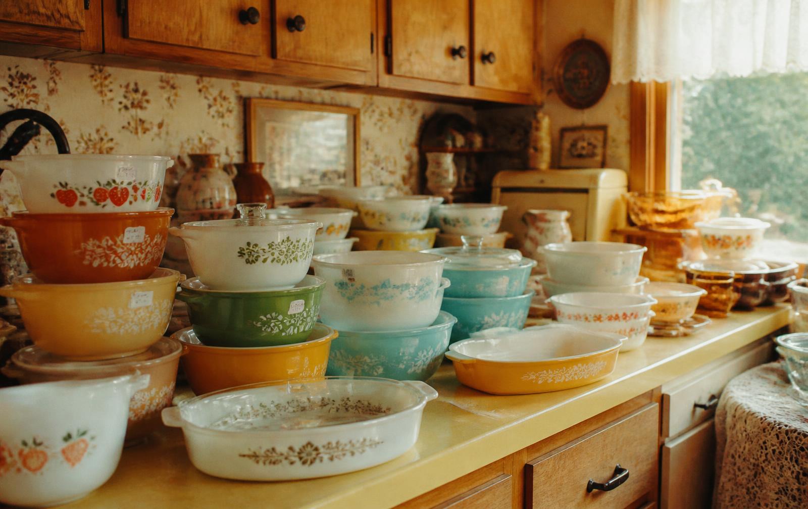 A 1970s Midwest kitchen at an estate sale, counters crowded with stacks of colorful patterned vintage Pyrex bowls and casseroles with handwritten price stickers.