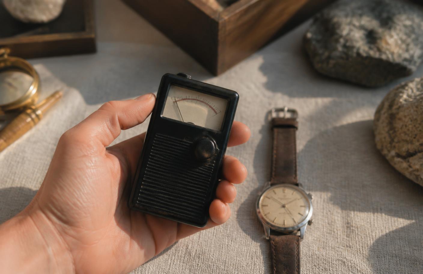 A collector holding a hand-held Geiger counter over the dial of a vintage wristwatch on a workbench