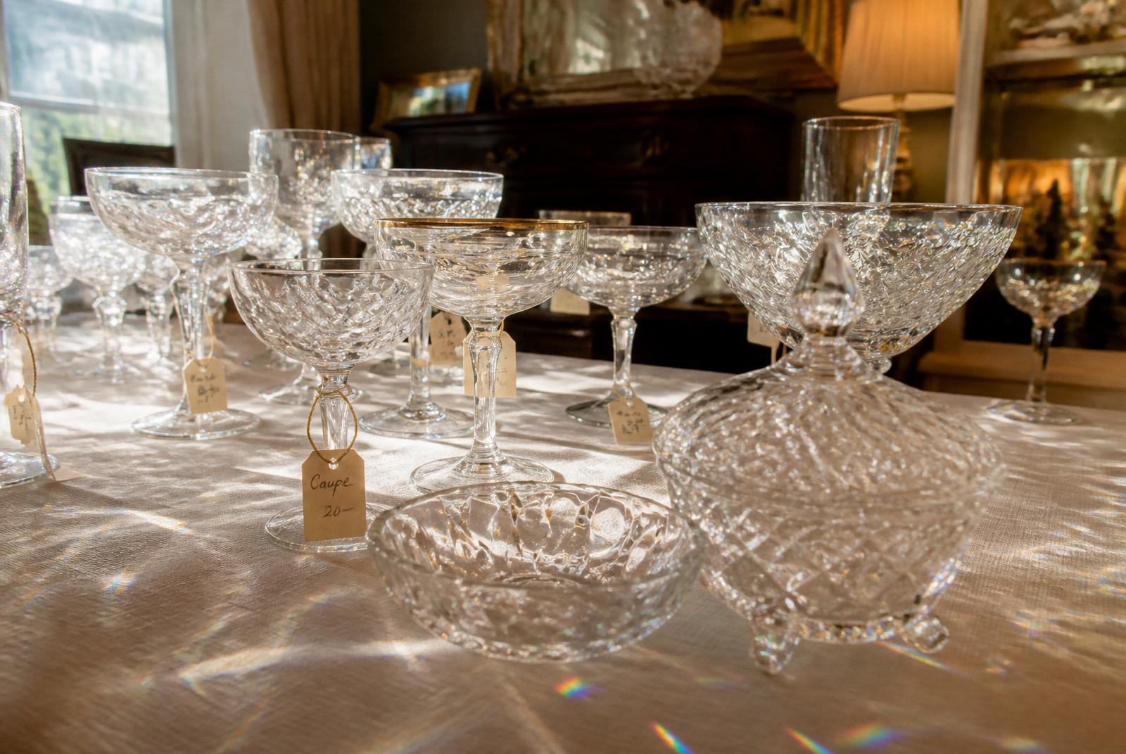 A collection of mismatched vintage champagne coupes and cut-glass crystal on an estate sale table, sunlight casting rainbow prisms across a lace tablecloth