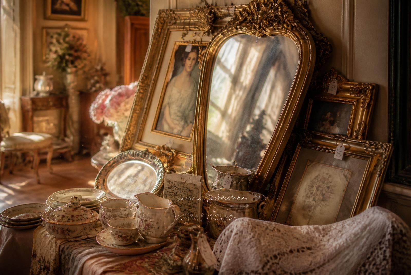 Ornate vintage gold picture frames and an antique mirror leaning against a wall at an estate sale, beside vintage china, silver trays, and lace linens in warm sunlight