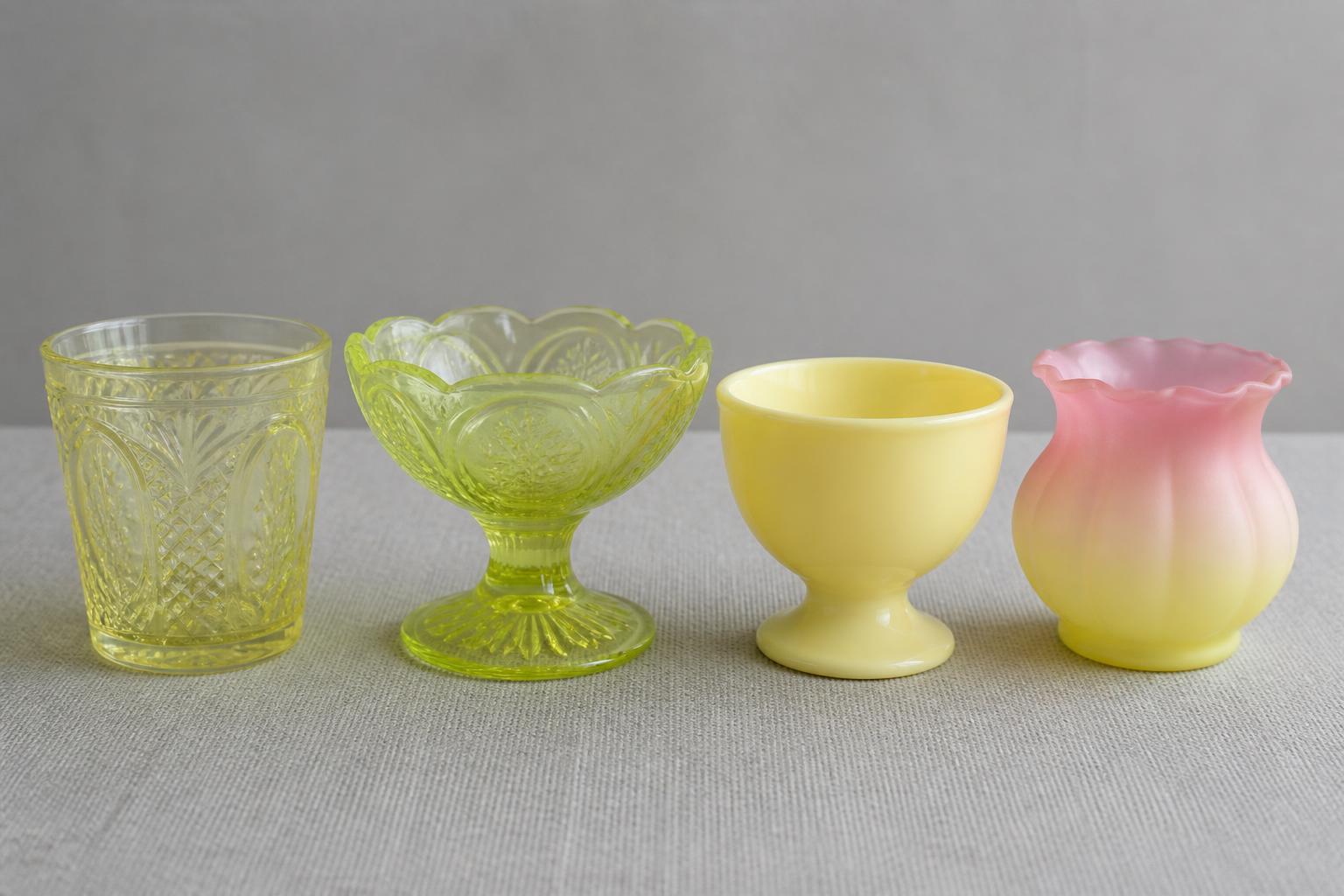 Four antique glass pieces in a row on grey linen: a transparent pale yellow canary tumbler, a yellow-green vaseline compote, an opaque creamy custard cup, and a satin piece shading pink at the rim to yellow at the base