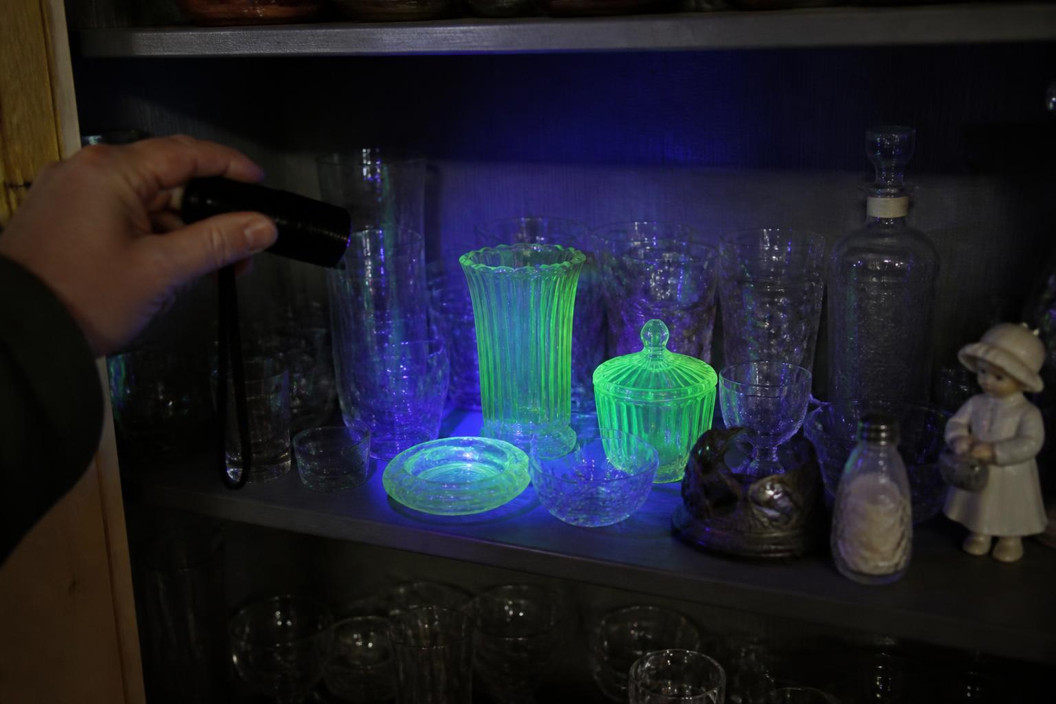 A shopper sweeping a small ultraviolet flashlight across a crowded dim shelf of vintage glassware at an estate sale, where two pieces light up vivid green while the rest stay dull