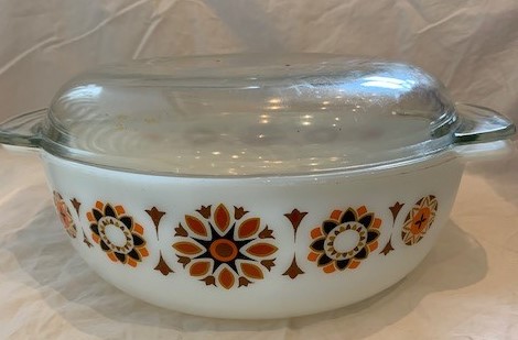 A round vintage Pyrex casserole with lid in a brown atomic-flower pattern — the tab-handled style that dominated mid-century kitchens.