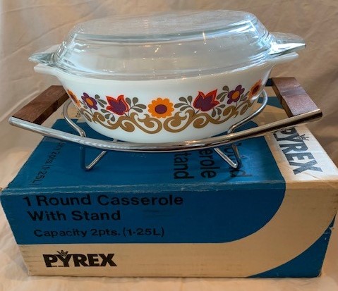 A patterned vintage Pyrex casserole sitting on its original "PYREX — 1 Round Casserole With Stand" box, showing the all-caps logo of the promotional era.
