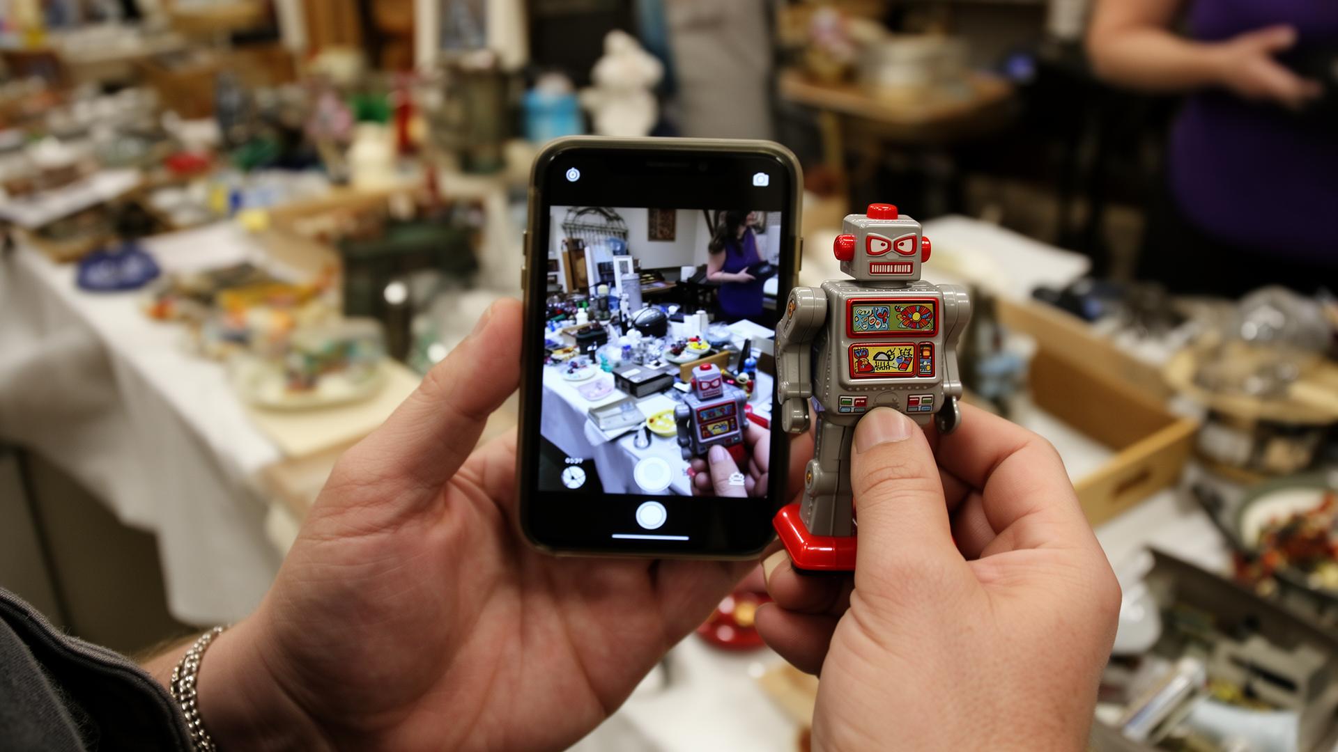 A person's hands at an estate sale photographing a small vintage plastic toy robot with a smartphone to check its value.